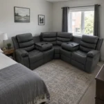 Charcoal corner recliner sofa in a simple guest bedroom with plain walls, a modest window, a small side table, and cool natural daylight.