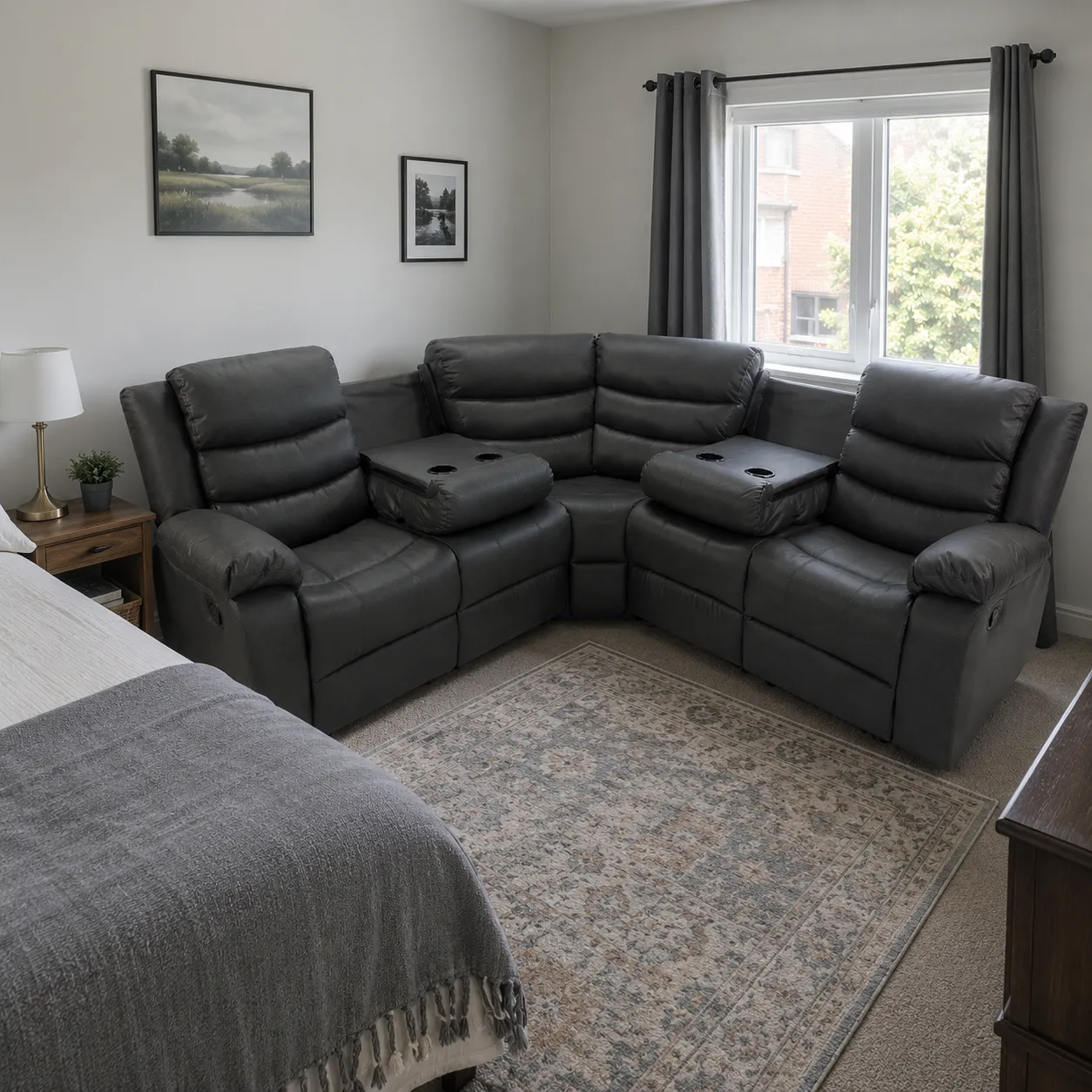 Charcoal corner recliner sofa in a simple guest bedroom with plain walls, a modest window, a small side table, and cool natural daylight.